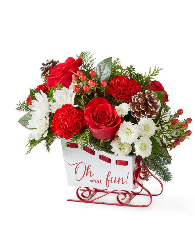 Festive holiday bouquet featuring red roses, red carnations, white button poms, and white cushion poms accented with lush seasonal greenery. Arranged in a cheerful container, this classic red and white floral display captures the joyful spirit of the seas