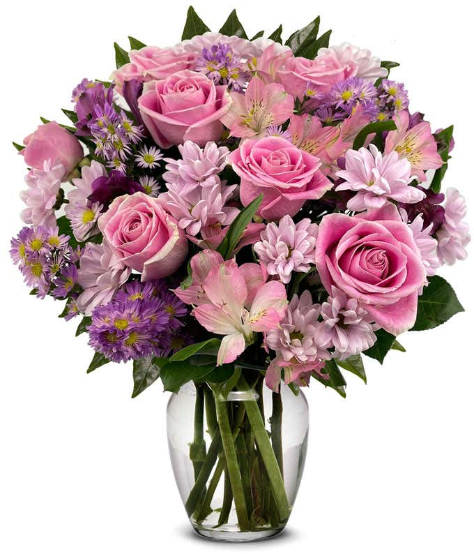 A full bouquet of pink roses, lavender daisy poms, pink alstroemeria, and purple asters in a clear glass vase with leafy greenery.