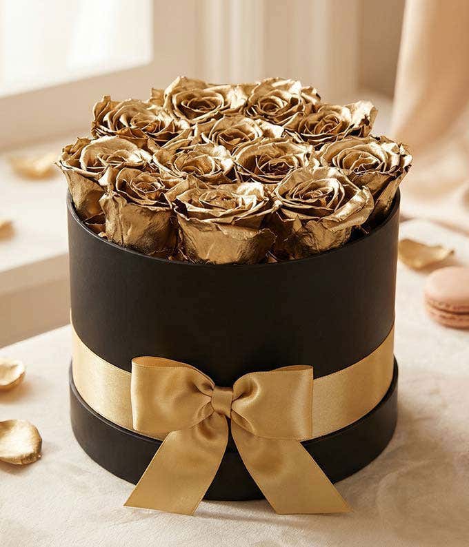 One dozen gold preserved roses arranged in a black hat box with a gold ribbon.