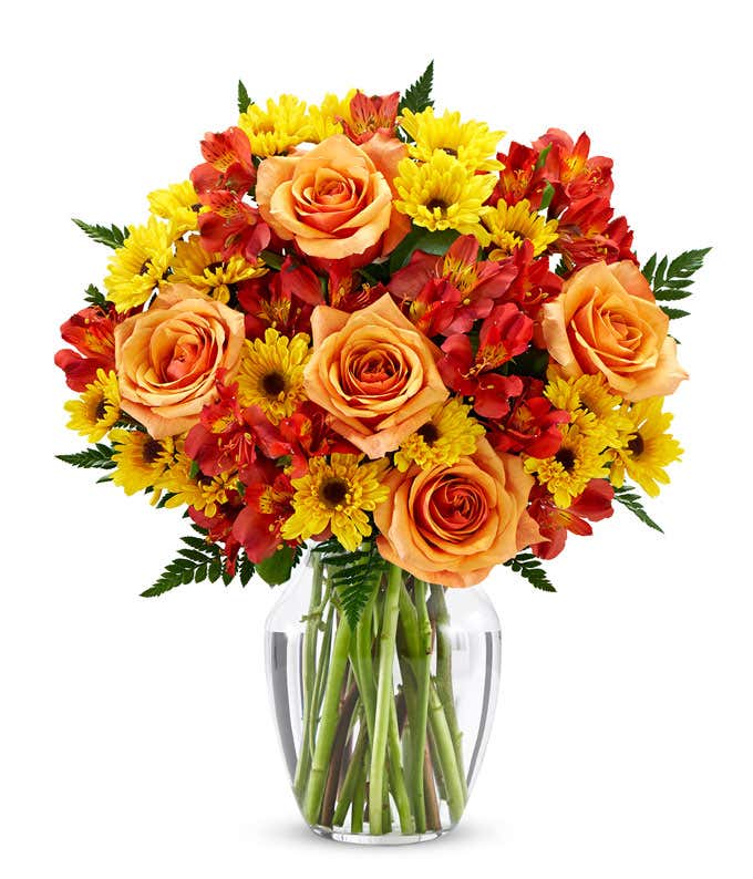 A bright fall bouquet of orange roses, red alstroemeria, and yellow daisies with greens in a glass vase.