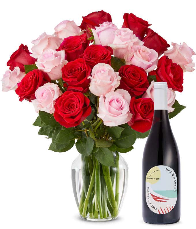 A mix bouquet of two dozen red and blush roses fills a clear glass vase and is paired with a bottle of Hill & Harbor California Pinot Noir.