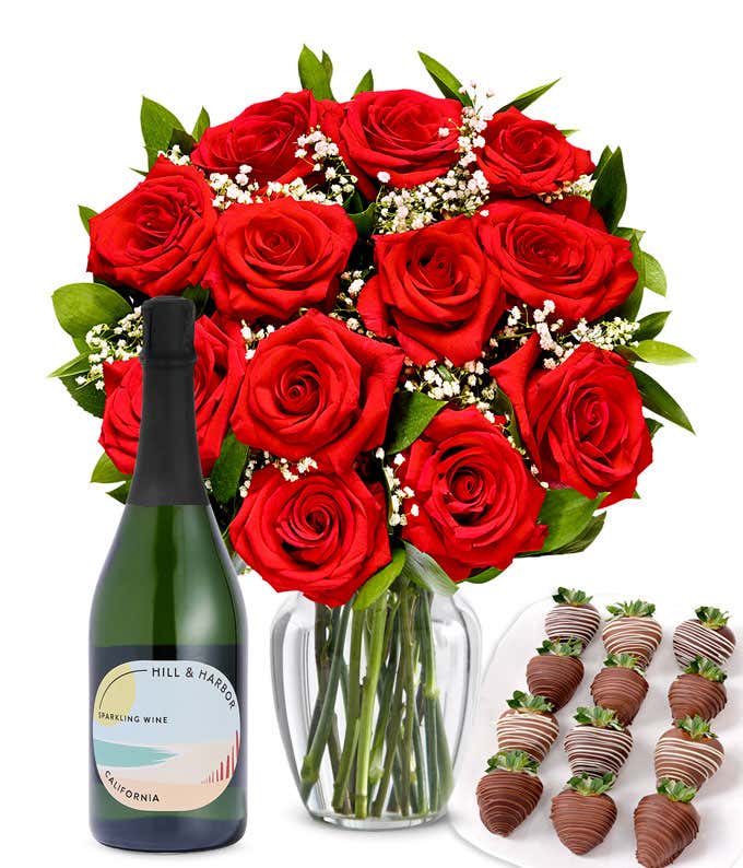 A bouquet of 1 dozen red roses in a clear glass vase is displayed beside a bottle of Hill & Harbor Sparkling Wine and an arrangement of 12 chocolate-covered strawberries.