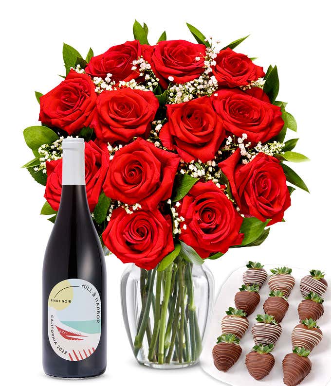 A bouquet of 1 dozen red roses in a clear glass vase is displayed beside a bottle of Hill & Harbor Pinot Noir and an arrangement of 12 chocolate-covered strawberries.