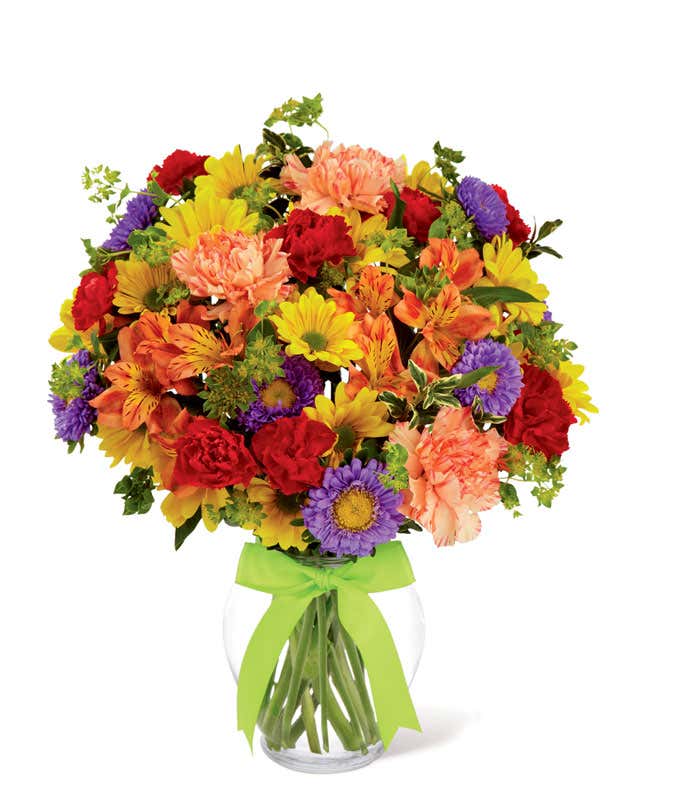 A full and colorful arrangement of small flowers, with shades of red, orange, yellow, purple, and peach, in a glass vase tied with a green ribbon