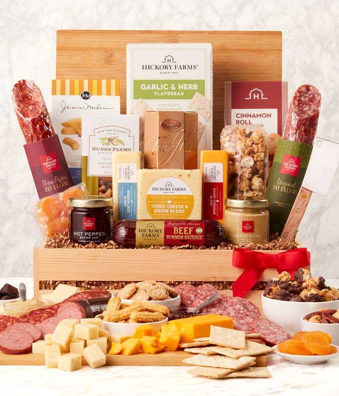 Gourmet charcuterie gift box with assorted salami, beef and turkey summer sausages, Three Cheese & Onion Blend, crackers, garlic & herb flatbread, hot pepper bacon jam, mustard, nuts, almond biscotti, and olives, arranged with a red bow.