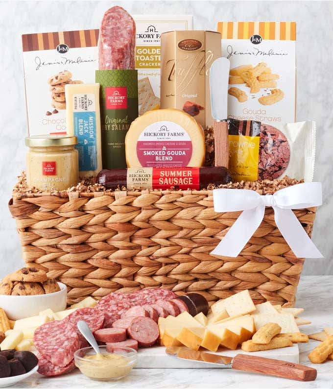 Woven gift basket with salami, cheese, mustard, crackers, cookies, and chocolates. In front, a board displays sliced meats, cheese cubes, and mustard for dipping, with cookies and chocolate truffles arranged alongside. Decorated with a white bow.