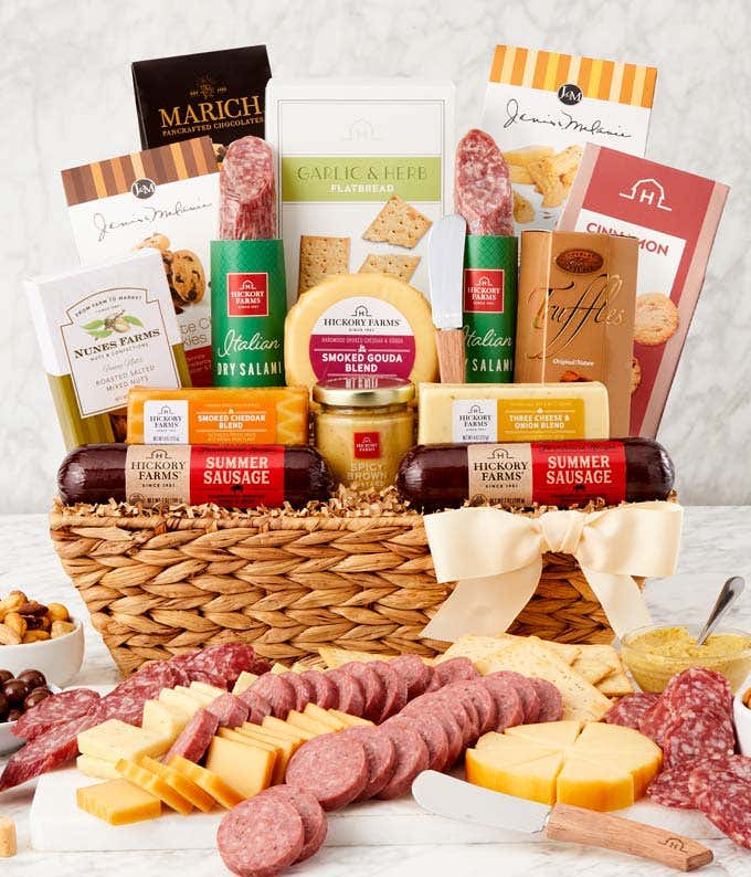 Woven gift basket filled with summer sausages, salami, assorted cheeses, crackers, mustards, cookies, chocolate truffles, and snacks. A serving board in front displays sliced sausage, cheese wedges, mustard, and pretzels, with a white bow on the basket.