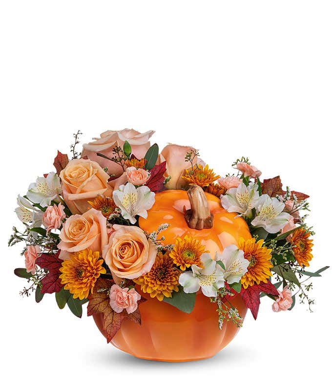 Floral arrangement of peach roses, white alstroemeria, miniature peach carnations, and bronze cushion spray chrysanthemums with greenery, displayed in a bright orange pumpkin-shaped container.