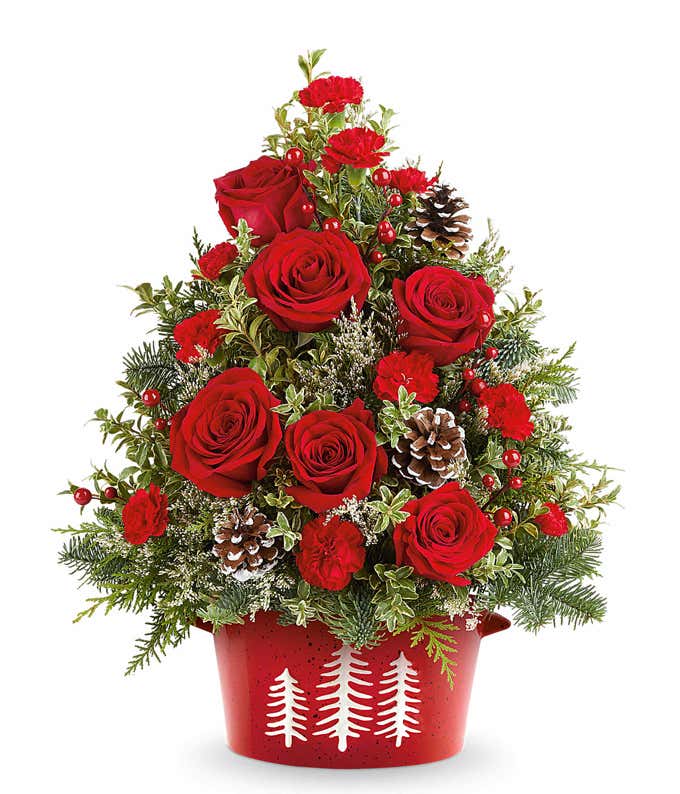Christmas tree-shaped floral arrangement with red roses, red carnations, pinecones, berries, and greenery in a red container decorated with white tree silhouettes.