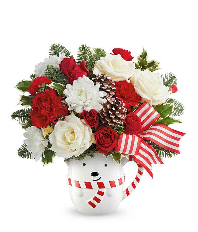 Festive holiday arrangement with red and white roses, red carnations, white mums, pinecones, greenery, and a red-and-white striped bow in a polar bear mug container.