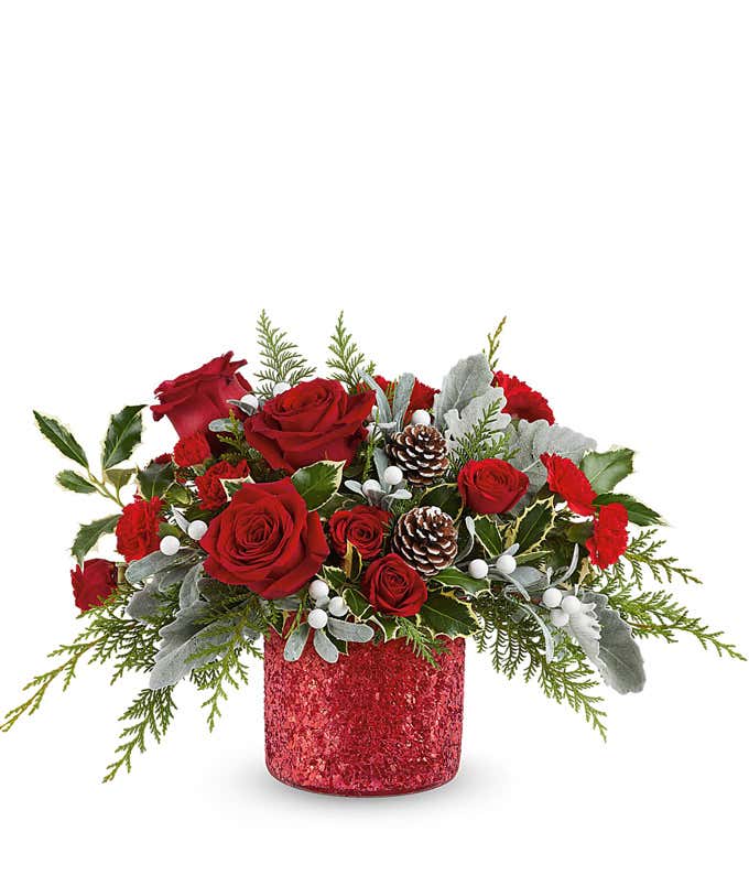 Holiday floral arrangement with red roses, red carnations, white berry picks, frosted pinecones, and seasonal greenery in a red glittered container.