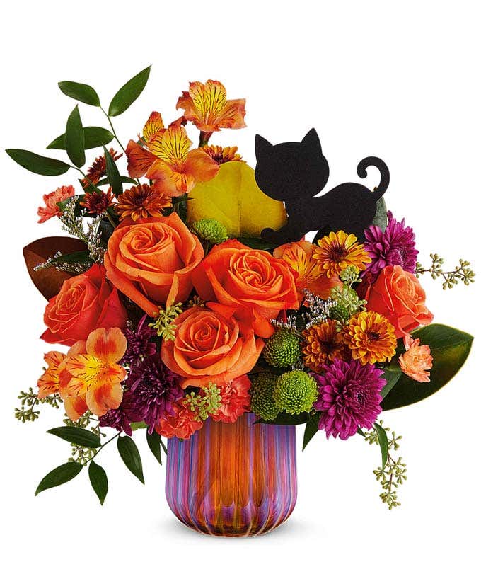 Orange Roses, orange alstroemeria, mini orange carnations, bronze poms, green poms, purple poms, and floral greenery in a purple glass vase with a black cat pick