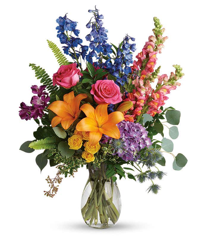 A vibrant arrangement featuring purple hydrangea, pink roses, yellow spray roses, orange Asiatic lilies, purple alstroemeria, blue delphinium, pink snapdragons, blue eryngium, huckleberry, eucalyptus, and fresh greenery, beautifully presented in a clear g