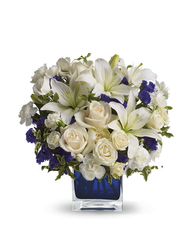 Floral arrangement featuring crème roses, crème spray roses, white Asiatic lilies, white miniature carnations, and purple statice in a blue cube vase.