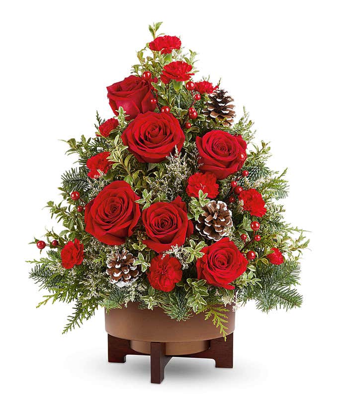 Christmas tree-shaped floral arrangement featuring red roses, red carnations, frosted pinecones, red berry accents, and assorted greenery in a brown wooden-style container.