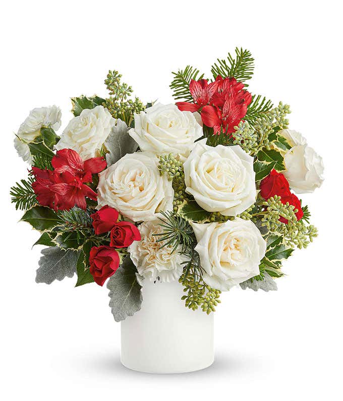 White roses, red alstroemeria, red spray roses, white carnations, and holiday greenery arranged in a round white vase.