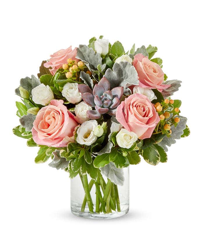 Floral arrangement in a clear glass vase featuring soft pink roses, white spray roses, peach hypericum berries, silver dusty miller, green foliage, and a central mauve-toned succulent.