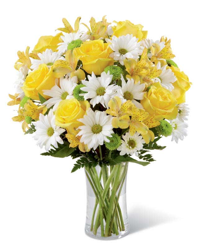 Bright bouquet featuring yellow roses, yellow alstroemeria, white daisies, green poms, and assorted greenery arranged in a tall clear glass vase. 