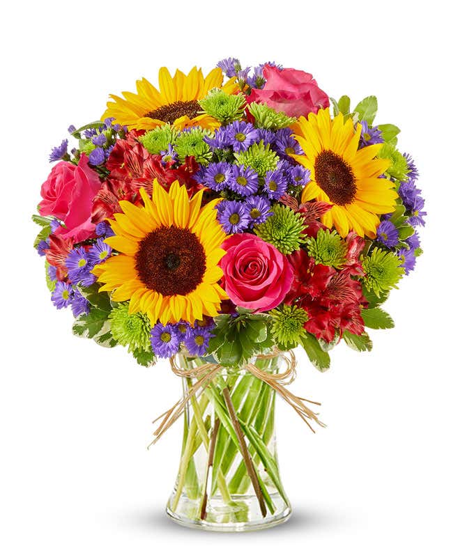 Bright mixed flower bouquet featuring yellow sunflowers, hot pink roses, red alstroemeria, green button poms, and purple asters in a clear glass vase tied with raffia ribbon.