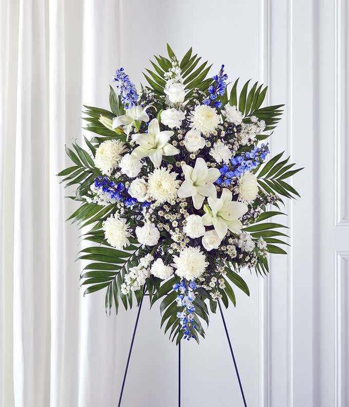 Standing spray arrangement featuring white lilies, roses, carnations, and poms, accented with blue delphinium, white monte casino, and palm fronds on a display easel. 