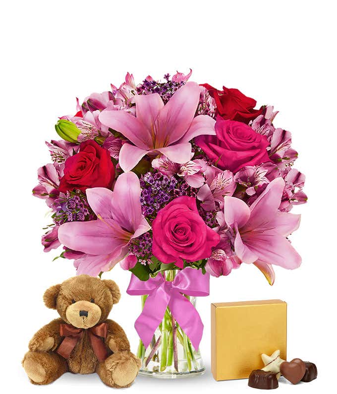 Romantic gift set with a bouquet of pink lilies, hot pink and red roses, pink alstroemeria, and purple filler in a glass vase with a pink bow, paired with a plush teddy bear and box of assorted chocolates. 