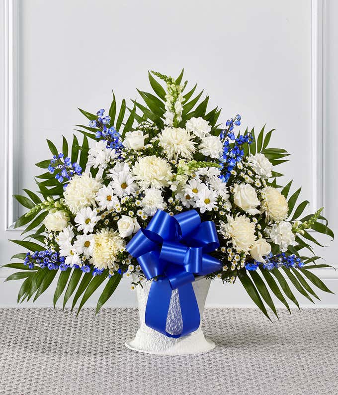 Standing floral arrangement of white roses, chrysanthemums, carnations, daisies, and snapdragons accented with blue delphinium and a large royal blue ribbon bow.