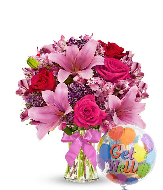 Clear glass vase filled with pink lilies, hot pink roses, red roses, pink alstroemeria, and purple filler flowers, tied with a pink bow and paired with a colorful 'Get Well' balloon. 