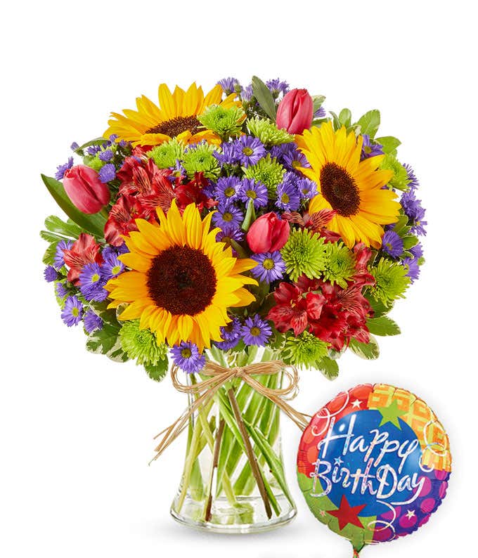 Clear glass vase filled with sunflowers, red alstroemeria, pink tulips, green button poms, and purple asters, tied with raffia and paired with a colorful Happy Birthday balloon.