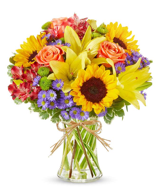 An arrangement of sunflowers, yellow lilies, orange roses, alstroemeria, Monte Casino, and green poms, in a clear glass vase.