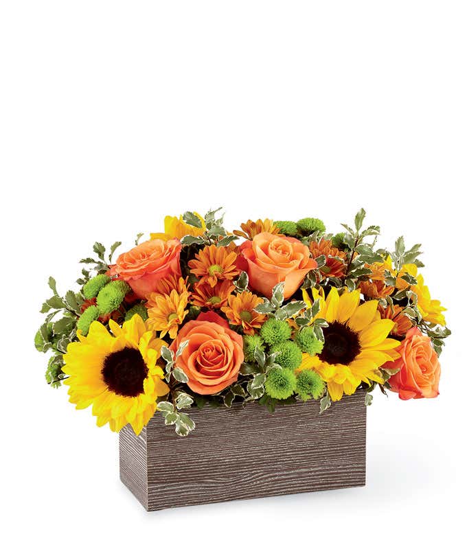Fall flower arrangement with orange roses, yellow sunflowers, bronze daisies, green button poms, and variegated greenery in a rectangular wood-textured container.
