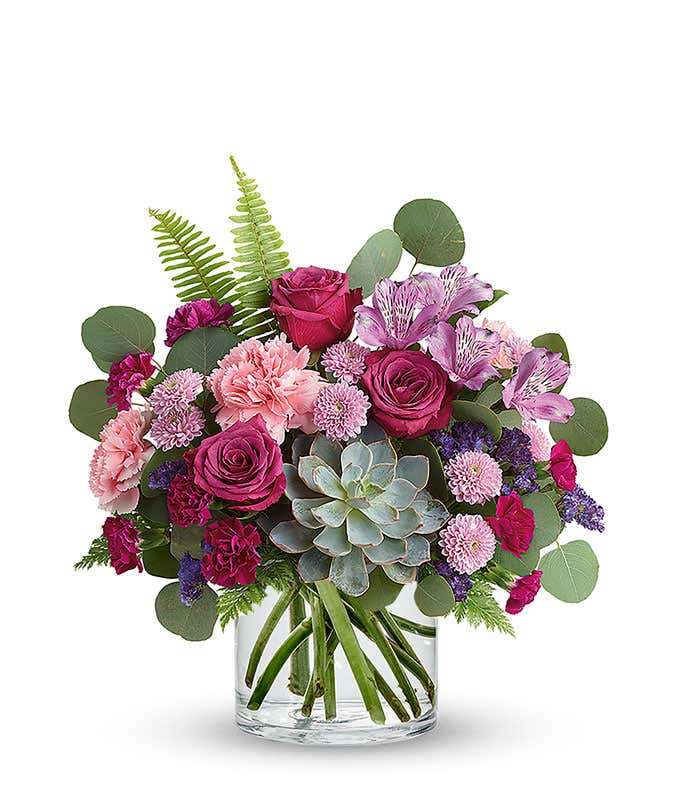 Floral arrangement featuring a succulent plant, pink carnations, purple mini carnations, dark pink roses (in deluxe and premium versions), purple statice, purple alstroemeria, lavender button mums, seasonal greenery in a glass vase