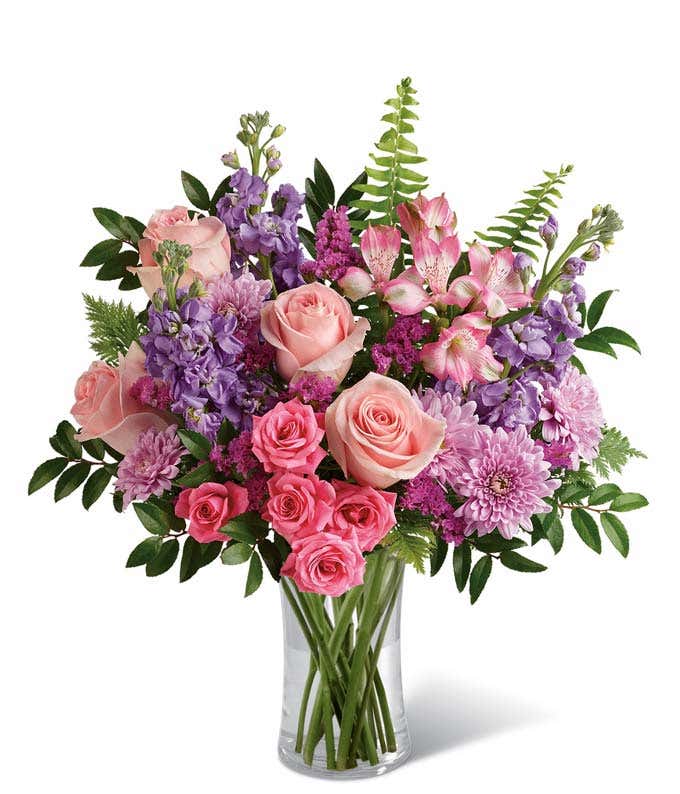 Floral arrangement featuring light pink roses, pink spray roses, pink alstroemeria, lavender stock, lavender mums, raspberry statice, and greenery in a glass vase.