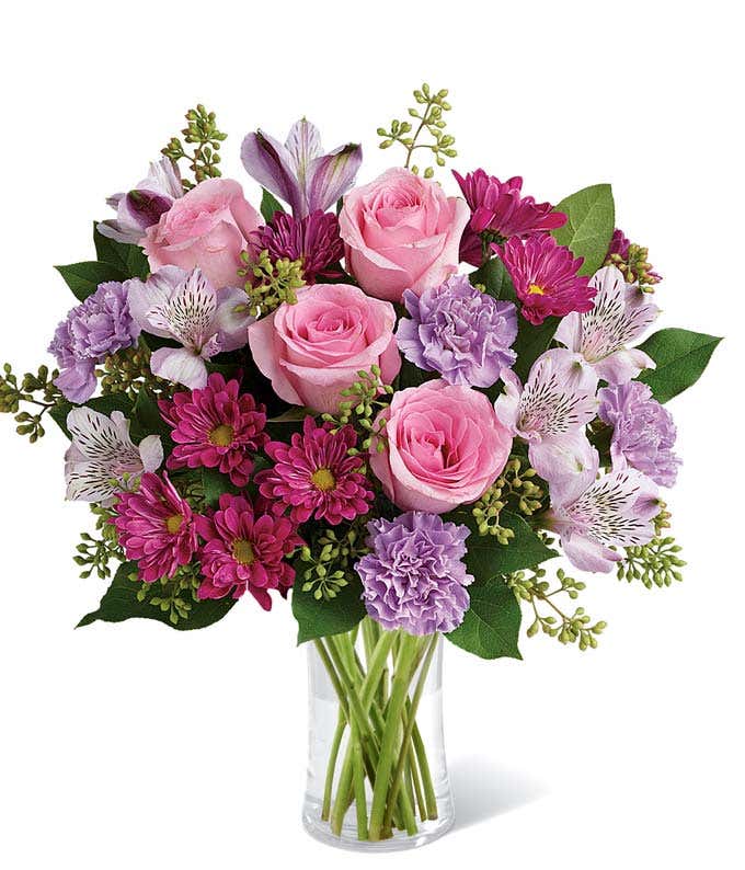 Pink roses, lavender carnations, magenta daisies, white alstroemeria, and lush greenery arranged in a clear glass vase, creating a soft and elegant bouquet in shades of pink and purple.