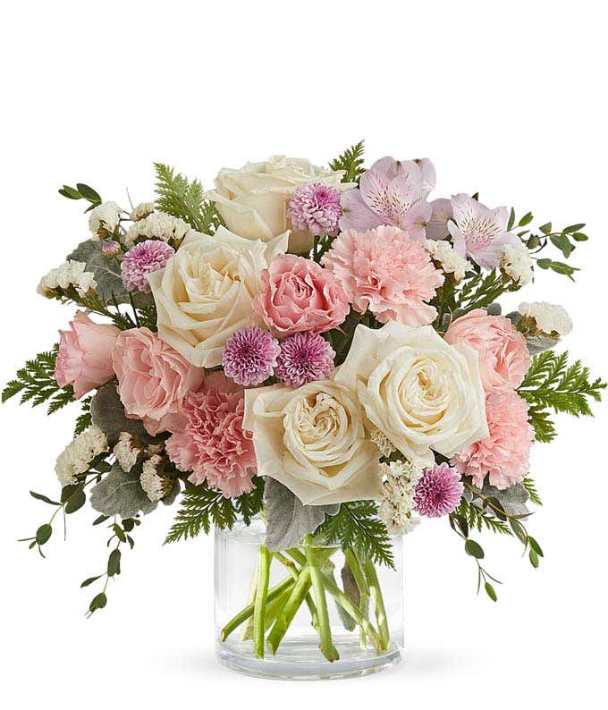 A bouquet of White Roses, Pink Spray Roses, Lavender Alstroemeria, Pink Carnations, Lavender Button Poms, and Floral Greenery in a short clear cylinder vase