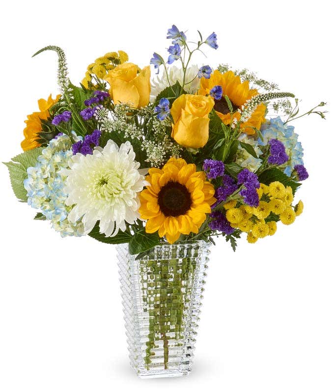 A bouquet of yellow roses, buttons, and sunflowers, blue hydrangea and delphinium, white mums, stock, and queen anne's lace and purple statice in a clear glass vase.