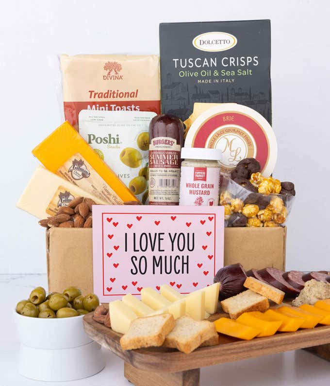 A vibrant gift box featuring 3 cheeses, gourmet crackers, focaccia crisps, summer sausage, olives, smoked almonds, stone-ground mustard, chocolate-covered popcorn, and a pink card with hearts reading 'I Love You So Much.'