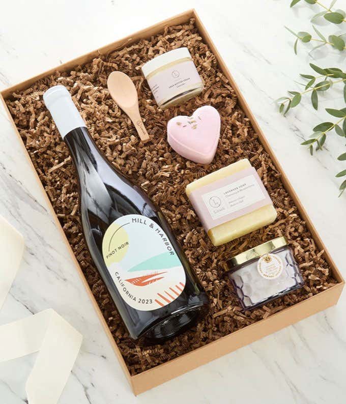 Bottle of Hill & Harbor Pinot Noir displayed with spa essentials including artisan soap, bath salts with wooden scoop, and a scented candle arranged on a white surface, creating an elegant wine and self-care gift set.