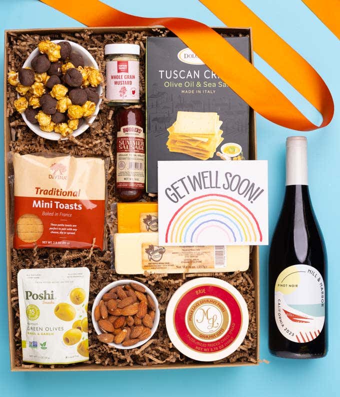 A charcuterie gift set with Tuscan crisps, focaccia crackers, 3 cheeses, summer sausage, olives, mustard, smoked almonds, chocolate-covered popcorn, a bottle of Pinot Noir, and a get well soon card.
