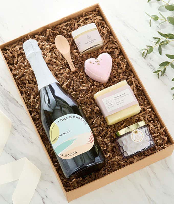 Bottle of Hill & Harbor Sparkling Wine displayed with spa essentials including artisan soap, bath salts with wooden scoop, and a scented candle arranged on a white surface, creating an elegant wine and self-care gift set.