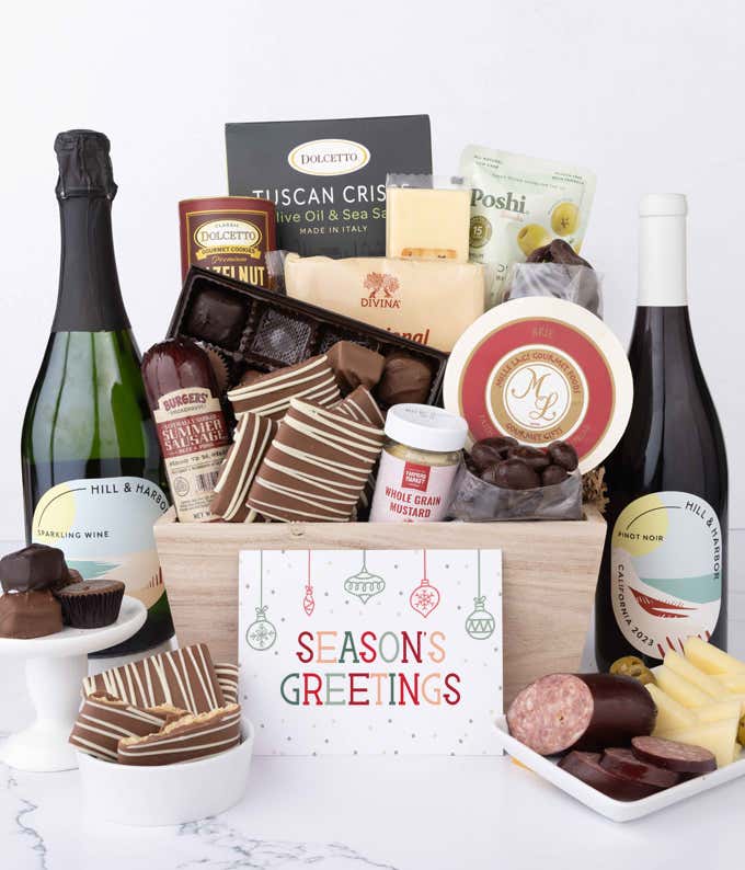 A wooden basket filled with chocolates, summer sausage, cheeses, crackers, crisps, mustard, and snacks, with displays of chocolate treats, sliced sausage, olives, and cheese arranged with 2 bottles of wine and Season's Greetings message.