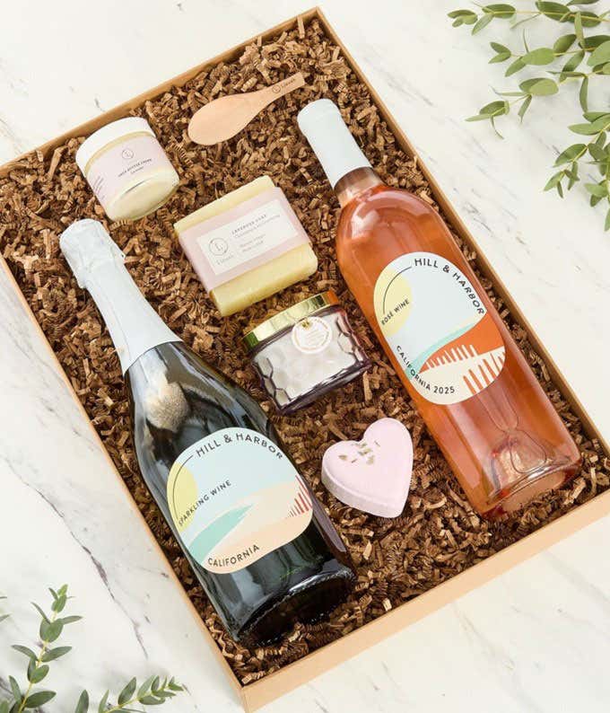 Two bottles of Hill & Harbor wines displayed with spa essentials including artisan soap, bath salts with wooden scoop, and a scented candle arranged on a white surface, creating an elegant wine and self-care gift set.