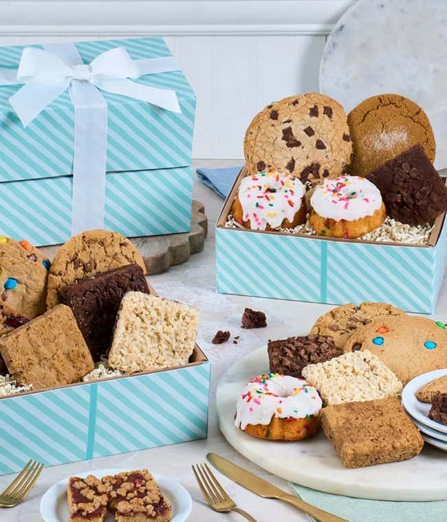 Two rainbow striped gift boxes with a blue bow. In front, a plate with a mini bundt cake with a candle, another tray with cookies, brownies, and blondies. Behind those, a cake tray with whoopie pies and mini bundt cakes. 