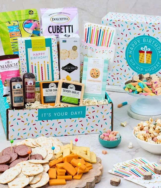 In a gift box, a bag of of chocolate wafter Cubetti's, a box of crackers, a bag of gummy worms, a box of kettle corn, three blocks of cheese, two packages of sausage. In front of the box, a plate of sliced meat &amp; cheese and a plate of crackers. Beside the