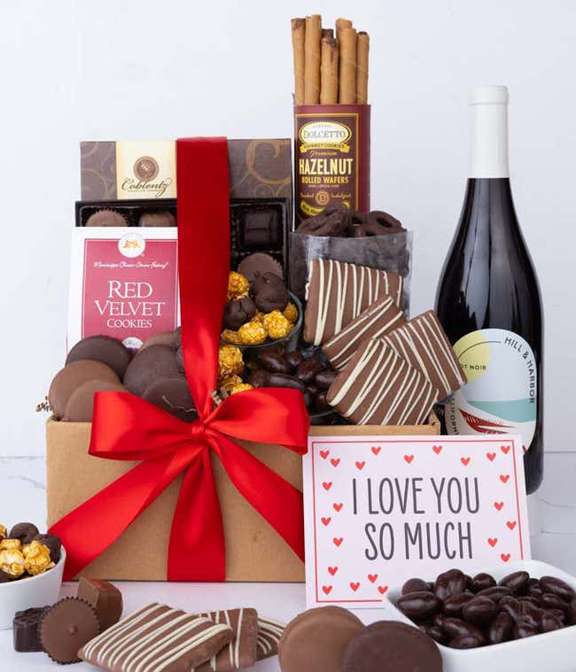 A gift box overflowing with treats including an assortment of chocolate-covered items like oreoes, almonds, graham crackers and pretzels along with red velvet cookies, hazelnut wafers, an 8 piece box of chocolates, a bottle of red wine and an I Love You c