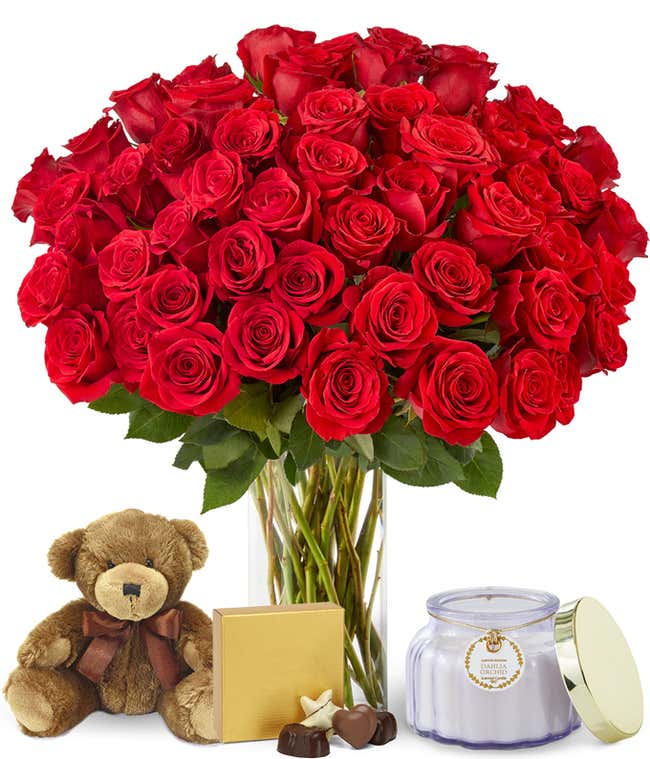 Stunning bouquet of numerous red roses, tightly arranged in a clear glass vase, creating a rich and luxurious floral display. Accompanied by a brown teddy bear, square box of chocolates, and purple candle.