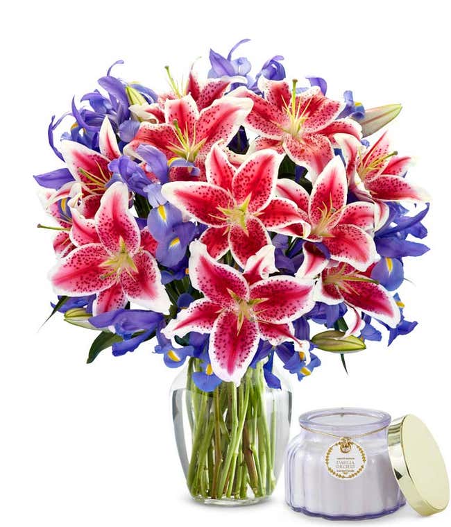 A bouquet of pink speckled lilies and blue/purple iris with yellow centers in a glass vase sitting next to a small purple candle.