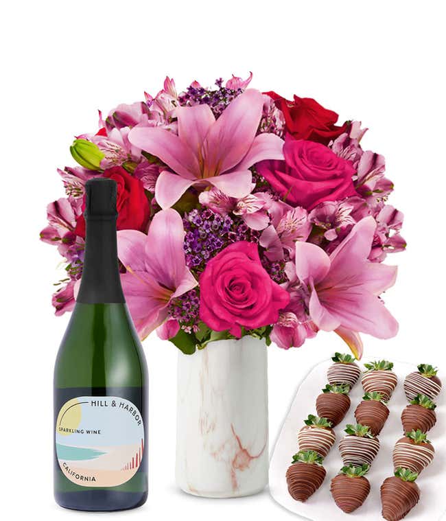 A mixed bouquet of pink and purple flowers in a clear glass vase is displayed with a bottle Hill &amp; Harbor Sparkling Wine and 12 chocolate-covered strawberries.