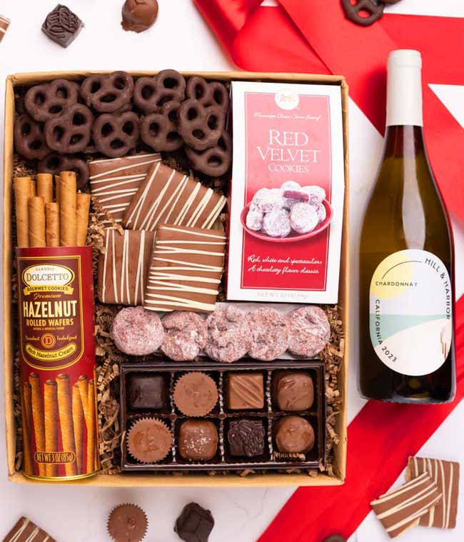 A gift box with chocolate pretzels, chocolate graham crackers, red velvet cookies, hazelnut wafer rolls, an 8 piece box of chocolates and a bottle of white wine.