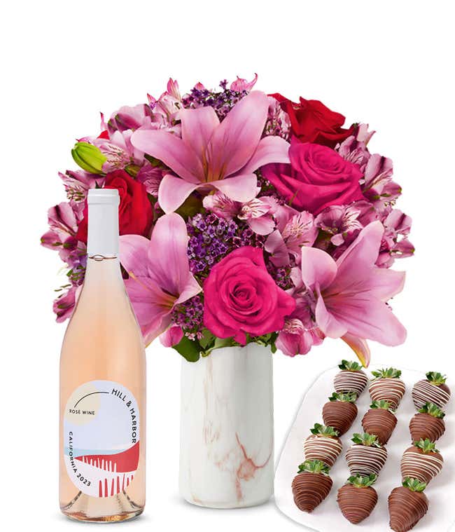 A mixed bouquet of pink and purple flowers in a clear glass vase is displayed with a bottle Hill &amp; Harbor Ros&eacute; and 12 chocolate-covered strawberries.