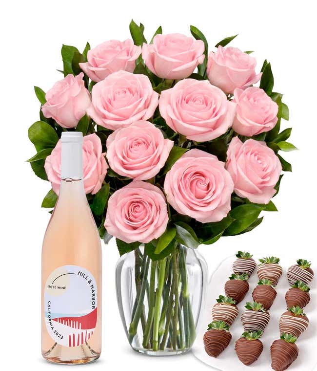 A bouquet of 1 dozen soft pink roses arranged in a clear glass vase is displayed beside a bottle of Hill &amp; Harbor Ros&eacute; and a neat assortment of 12 chocolate-covered strawberries.